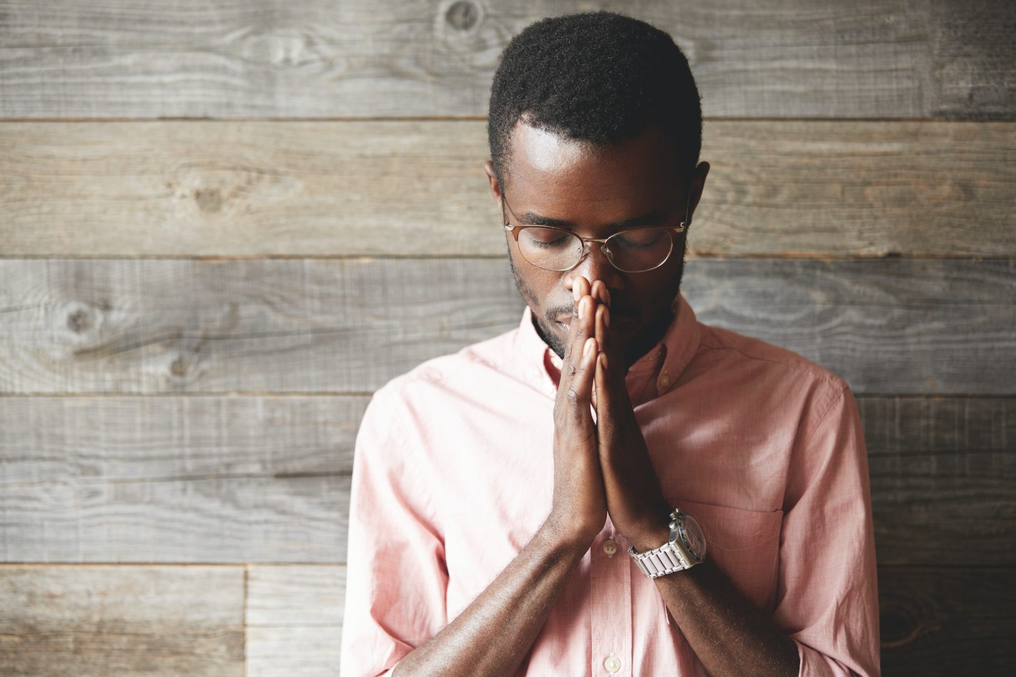 young-meditating-and-praying-african-american-man-wearing-pink-shirt-and-glasses-holding-hands-in-p-1980x1320